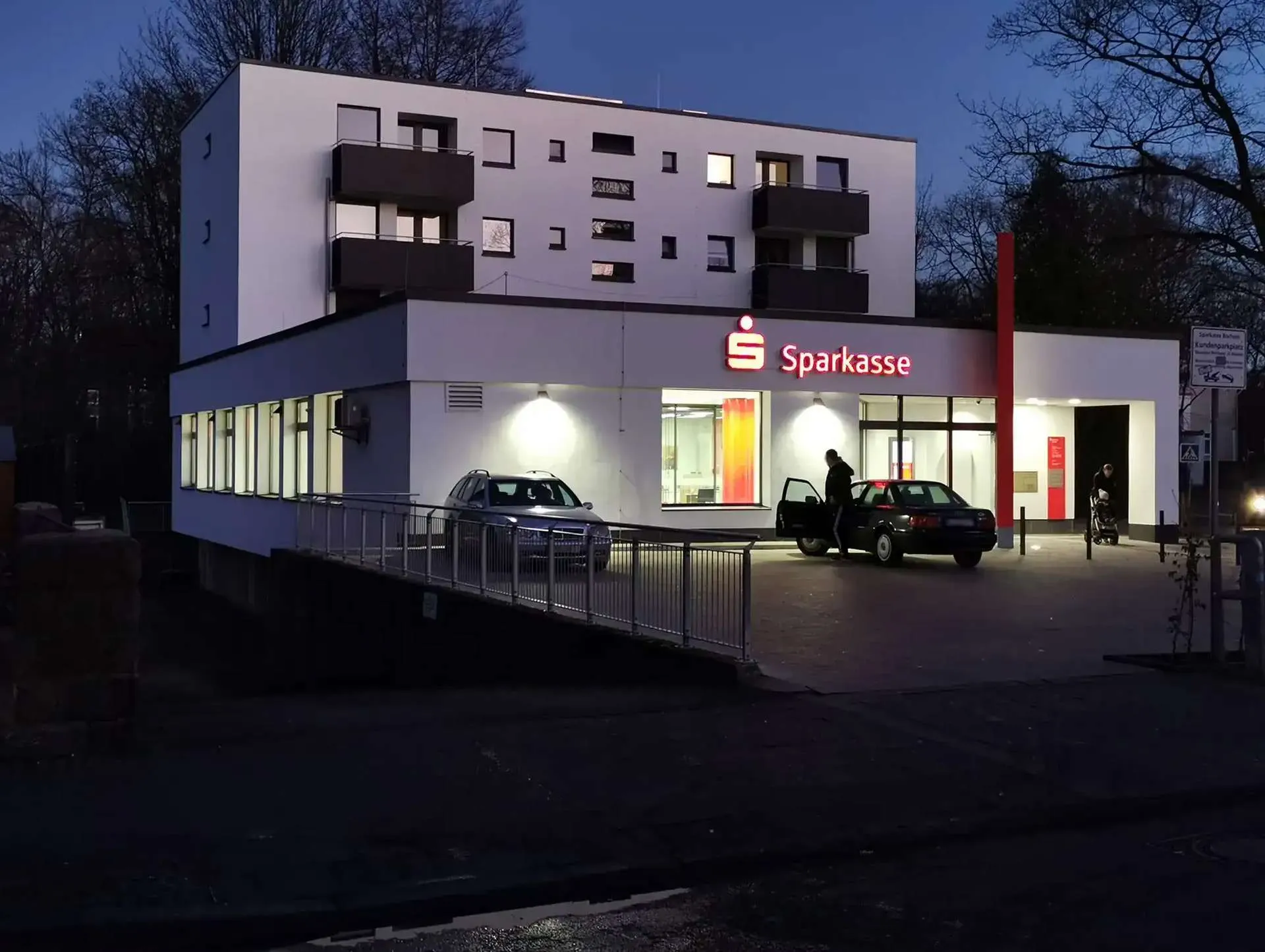 Beleuchtete Nord-West-Fassade der sanierten Sparkassen-Filiale mit Wohnhaus in Bochum bei Nacht, moderne Architektur, energetische Sanierung, farbige Akzente und freundliche Atmosphäre